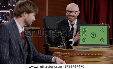Late-night talk show host showing a green board with tracking points to celebrity guest in a studio. TV broadcast style show