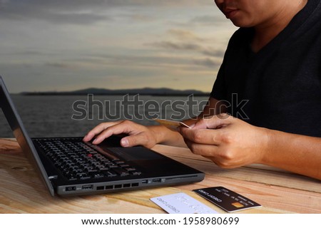 An Asian man sits on a brown wooden table, shopping online through a notebook and ordering through credit cards and overlooks the ocean.