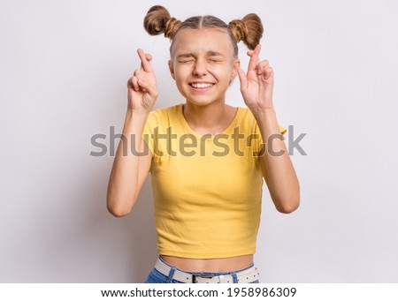 Portrait of funny teen girl crossing her fingers and wishing for good luck on gray background. Caucasian young teenager praying with crossed fingers and eyes closed. Child face expression emotions