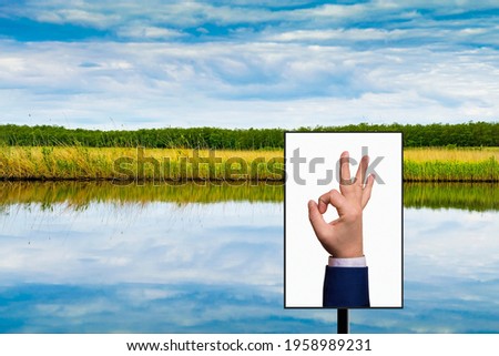 A man's hand shows an OK sign on a billboard against the background of a river, reeds, forest and blue sky. Mockup. Copy space