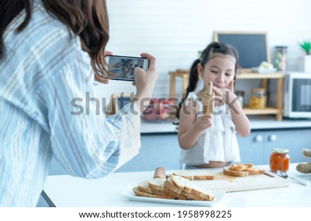 Mom taking pictures of her daughter or recording a vlog, making video call on phone having fun together in kitchen