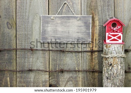 Blank rustic sign hanging over barbed wire fence post with red barn birdhouse