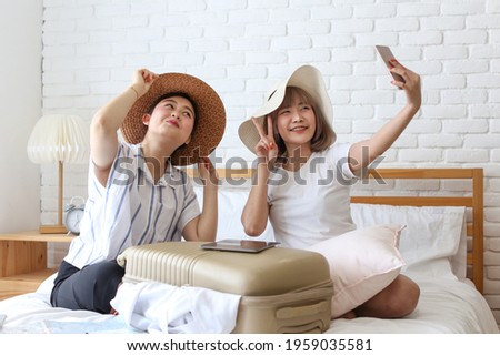 Young Asian couple taking selfie with mobile phone in apartment. Two women are preparing for the journey happily. Young female relaxing In room.