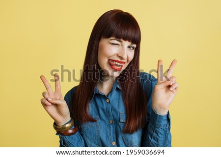 Funny happy laughing woman showing tongue, gesturing peace or victory sign and looking at the camera over yellow background. Close up studio shot.