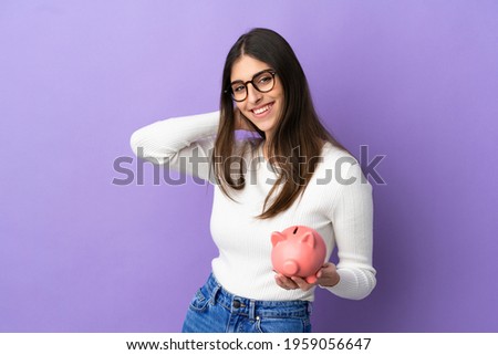 Young caucasian woman holding a piggybank isolated on purple background laughing