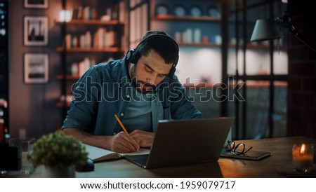 Handsome Caucasian Man With Headphones Listening Music on Laptop while Sitting in Dark Living Room in the Evening. Student Studying in Home School. Chat with Friends on Social Network. Royalty-Free Stock Photo #1959079717