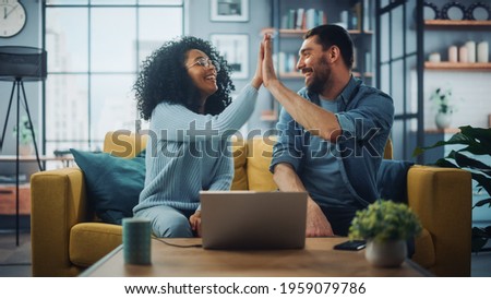 Diverse Multiethnic Couple are Sitting on a Couch Sofa in Stylish Living Room and Choosing Items to Buy Online with Laptop Computer, Give High Five. Friends or Colleagues are Discuss Work Projects. Royalty-Free Stock Photo #1959079786