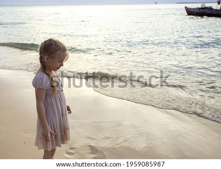 A blonde girl 4-5 years old stands on the white sand near the Indian Ocean in a light dress and looks down