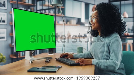 Latina Female Specialist Working on Desktop Computer with Green Screen Mock Up Display at Home Living Room while Sitting at a Table. Freelancer Female Chatting Over the Internet on Social Networks.