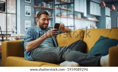 Happy Handsome Caucasian Man Using Smartphone in Cozy Living Room at Home. Man Resting on Comfortable Sofa. He's Browsing the Internet and Checking Videos on Social Networks and Having Fun. Royalty-Free Stock Photo #1959089296