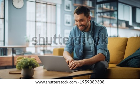 Handsome Caucasian Man Working on Laptop Computer while Sitting on a Sofa Couch in Stylish Cozy Living Room. Freelancer Working From Home. Browsing Internet, Using Social Networks, Having Fun. Royalty-Free Stock Photo #1959089308