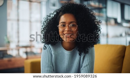 Portrait of a Beautiful Authentic Latina Female with Afro Hair Wearing Light Blue Jumper and Glasses. She Looks to the Camera and Smiling Charmingly. Successful Woman Resting in Bright Living Room. Royalty-Free Stock Photo #1959089326