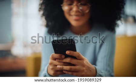 Portrait of a Beautiful Authentic Latina Female with Afro Hair in Stylish Cozy Living Room Using Smartphone at Home. She's Browsing the Internet and Checking Videos on Social Networks and Having Fun.