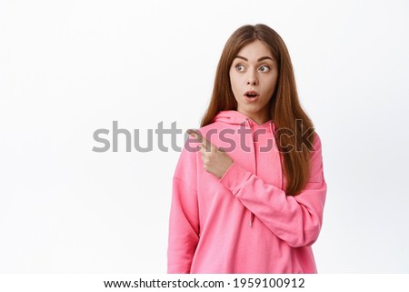 Surprised woman gasping, pointing and looking left aside, showing interesting thing, standing over white background.