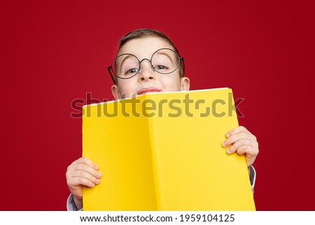 Clever little schoolboy in nerdy spectacles looking out from behind of bright yellow textbook and smiling against red background