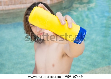 teen boy with long hair showing suntan lotion bottle while sitting in pool