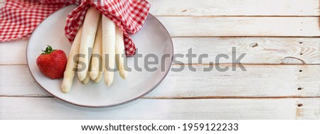 Uncooked white asparagus with strawbeery and red checkered cloth on vintage wooden planks. Fresh ingredients for a seasonal gastronomy background with space for text.