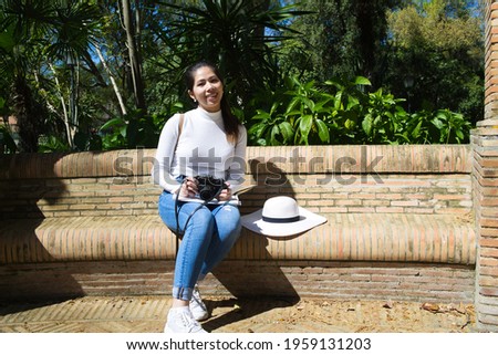 Asian woman on holiday in Spain. She is sitting in a park looking at the pictures on the camera while resting. On her legs she has a tourist map of the city. She is wearing a white hat to protect sun.
