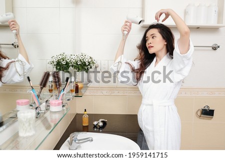 Attractive young Vietnamese woman blowing out hair in front of bathroom mirror