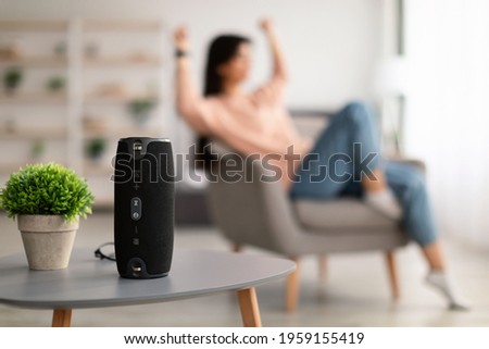 Modern Technology Concept. Closeup of smart portable wireless speaker on the table, selective focus. Young woman listening to music and dancing, sitting on armchair in the blurred background Royalty-Free Stock Photo #1959155419