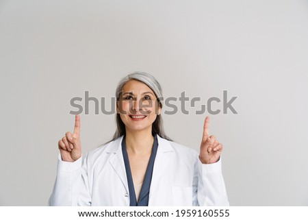 Asian mature woman in medical uniform smiling and pointing fingers upward isolated over white background
