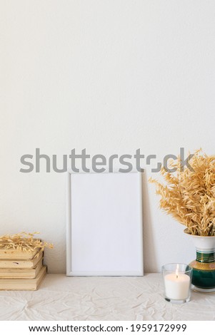Home decor mock-up, blank picture frame near white painted concrete wall , vase with dried yellow oat stalks, old books and a burning candle