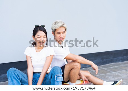 Half body of Asian handsome man and cute woman sitting on skateboards, look at right side, act smart, and smile in front of cool wall with copy space on wall in daylight time, summer holiday.