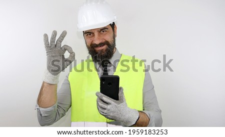 Architect wearing hardhat and reflecting jacket, smiling holding smatphone and making ok sign with hand. 