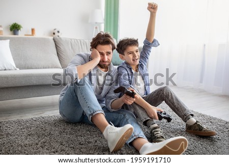 Video gamers. Joyful boy winning dad in online game, celebrating victory and raising hand. Father and son playing videogames, having fun at home, sitting on floor carpet, free space