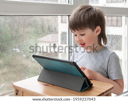 Curious boy watch cartoons on digital tablet. Kid uses electronic device. Indoor leisure for children while it's raining outside.