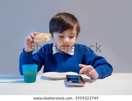 School kid eating homemade burger and typing on mobile phone, Hungry boy eating homemade bread sandwiches for his lunch, Child playig games on cellphone.
