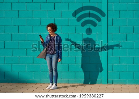 Front view of a happy woman standing against a bright blue wall using her smartphone with a shadow drawing a wifi signal Royalty-Free Stock Photo #1959380227