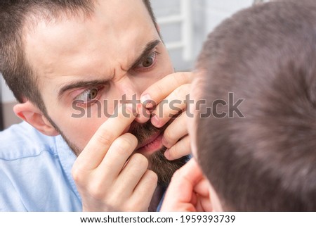 Bearded stares at a pimple on his nose, a man looks at himself in the bathroom mirror, portrait, close-up Royalty-Free Stock Photo #1959393739