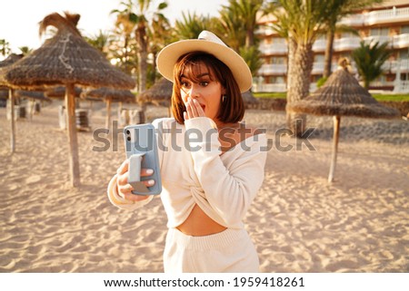 Summer beach vacation. Fashionable latino tourist woman taking fun mobile selfie photo with smartphone. Girl wearing hat, smiling and enjoying sunny day. Happy lifestyle concept. 
