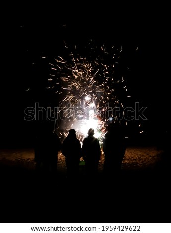 Group of people with back to camera in silhouette have a close up view of fireworks exploding on beach in black night.