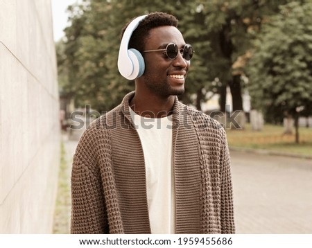 Portrait of happy smiling young african man in headphones listening to music wearing a brown knitted cardigan in a city