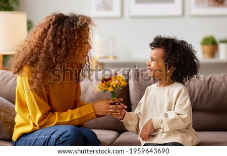 Excited boy little son congratulating his mom happy mixed race woman with Mothers day and giving her flower bouquet, they smiling while sitting on sofa at home. Family holidays concept