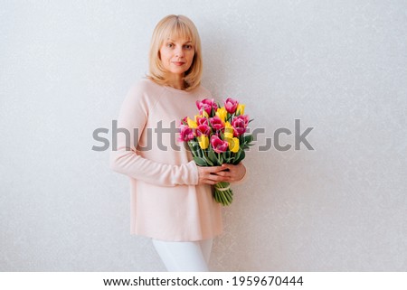 Portrait of Beautiful mature happy smiling woman with blonde hair, holding bouquet of pink and yellow tulip flowers in knitted bag. White background, copy space, close up.