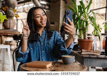 Black smiling woman gesturing peace sign while making video call in cafe