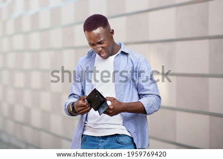 Desperate African American guy looking at his empty wallet near brick wall on city street. Unhappy black man with no cash in his purse outdoors. Economic crisis and bankruptcy concept Royalty-Free Stock Photo #1959769420