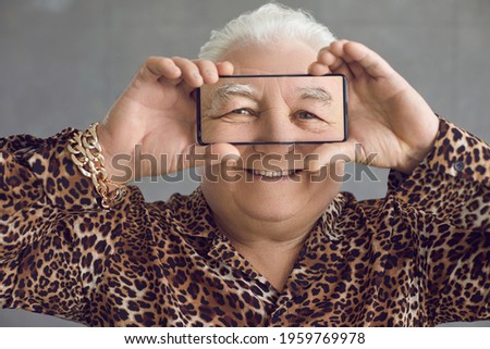 Funny chubby old guy having fun with mobile phone. Happy rich fat white haired senior man in trendy leopard print shirt and bling gold chain bracelet holding smartphone with zoomed image of his face