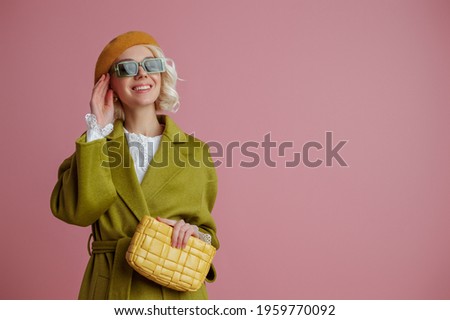 Spring fashion concept: happy smiling elegant lady wearing stylish green coat, sunglasses, beret, holding yellow leather padded bag, posing on pink background. Copy, empty space for text
