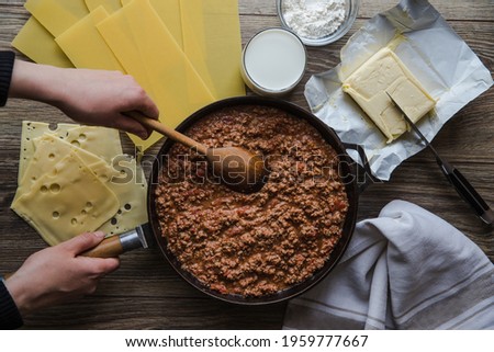 Woman cooking homemade classic lasagna bolognese, on dark table; with ingredients, top view copy space, hands in picture.