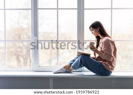Free Time To Relax. Profile of beautiful woman sitting on white windowsill at home, reading paper book, holding cup and drinking hot coffee. Happy lady taking break with mug near window, copy space Royalty-Free Stock Photo #1959786922