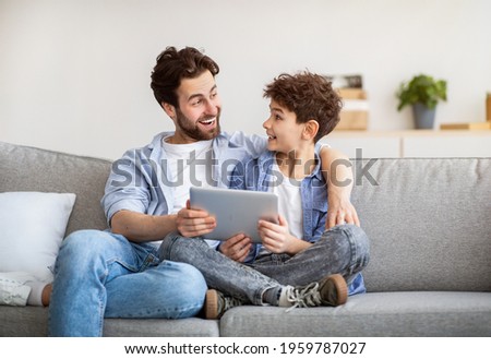 Family weekend. Excoted dad and son using tablet computer, surfing internet while spending time together at home, sitting on sofa in living room, copy space. Modern technologies concept