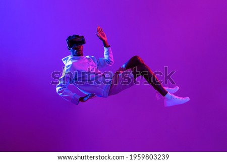 Amazed young African man wearing VR glass headset levitating in the air on futuristic purple cyberpunk neon light background