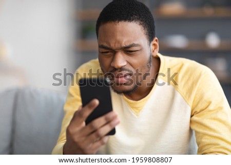 African american guy squinting while looking at mobile phone screen at home, copy space, blurred background. Young black man reading suspicious message on his smartphone, closeup Royalty-Free Stock Photo #1959808807