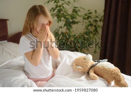 A little girl sits and sneezes on the bed among the blankets and pillows. House dust mite allergy Royalty-Free Stock Photo #1959827080