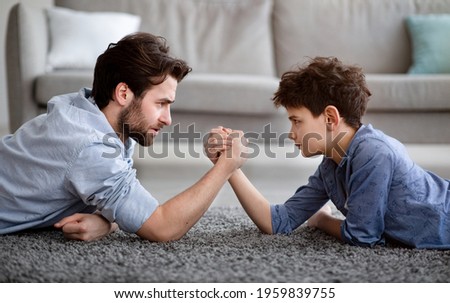 Raising a real man. Concentrated father and son arm wrestling and competing while lying on floor, having fun at home, side view. Family bond, leisure time and fatherhood concept