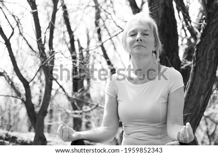 Mindful senior woman sitting in lotus position, meditating. Calm senior woman meditating in forest surrounded by trees. Black and white photo